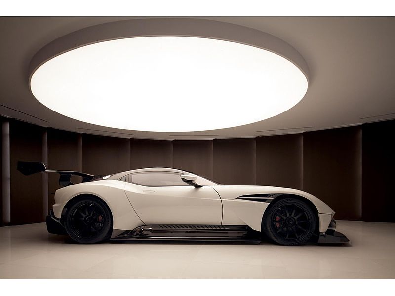 The image showcases a sleek, white sports car parked inside a modern garage. The car features black rims and a prominent rear spoiler, suggesting high performance. The garage is illuminated by a large, circular ceiling light, and the walls are lined with dark, vertical panels, creating a sophisticated and minimalist aesthetic.