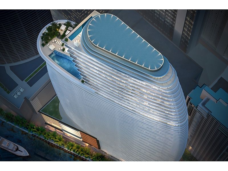 This aerial view showcases a modern, high-rise building with a unique, curved design. The building features multiple pools on its upper levels, surrounded by lounge areas and greenery. The overall impression is one of luxury and sophistication, highlighting the building's premium amenities and architectural appeal.