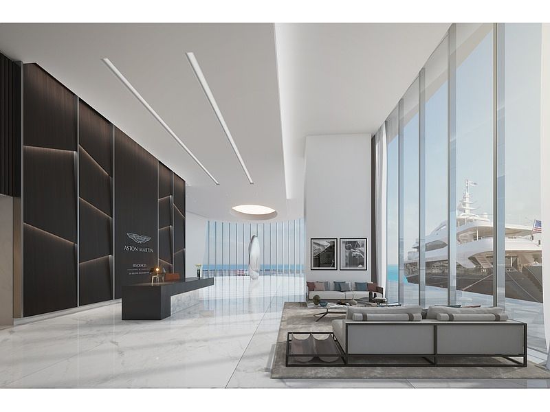This is a luxurious lobby area featuring a sleek reception desk with the Aston Martin logo, marble flooring, and floor-to-ceiling windows offering a view of yachts. The space is illuminated by modern linear lighting fixtures and includes a seating area with contemporary furniture, creating an upscale and welcoming atmosphere. The perspective is from the entrance, showcasing the depth and grandeur of the lobby.