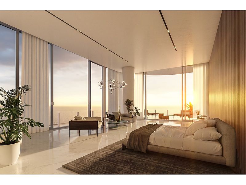 This is a luxurious primary bedroom with floor-to-ceiling windows offering stunning ocean views. The room features a neutral color palette, a large bed with plush bedding, and a modern chandelier. The overall impression is one of spaciousness, elegance, and tranquility, perfect for a high-end real estate listing.