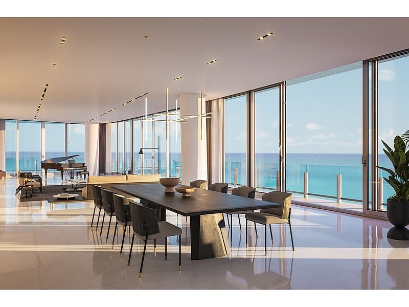 This is a luxurious dining room featuring a long, dark-colored dining table with modern chairs, illuminated by a contemporary chandelier. The room boasts floor-to-ceiling windows offering stunning ocean views, enhancing the sense of spaciousness and elegance. The marble flooring adds a touch of sophistication to the overall design.