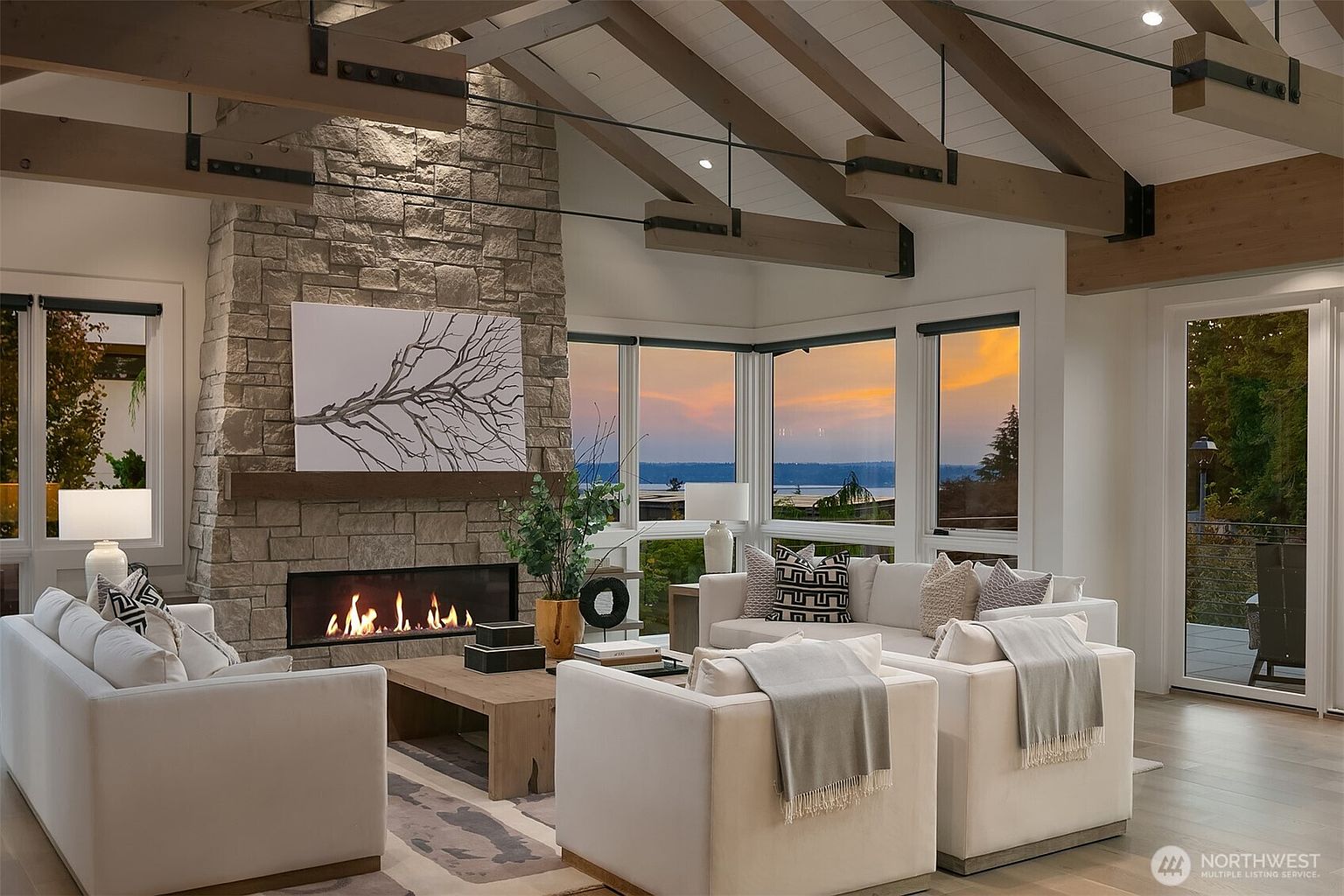 This sophisticated living room features a dramatic floor-to-ceiling stone fireplace as its centerpiece, complemented by exposed wooden ceiling beams and a vaulted ceiling. The space is furnished with plush, light-colored seating arranged around a wooden coffee table, all oriented toward expansive windows that offer a breathtaking sunset view over the water. The overall aesthetic is a blend of rustic warmth and modern luxury, creating an inviting and serene atmosphere.