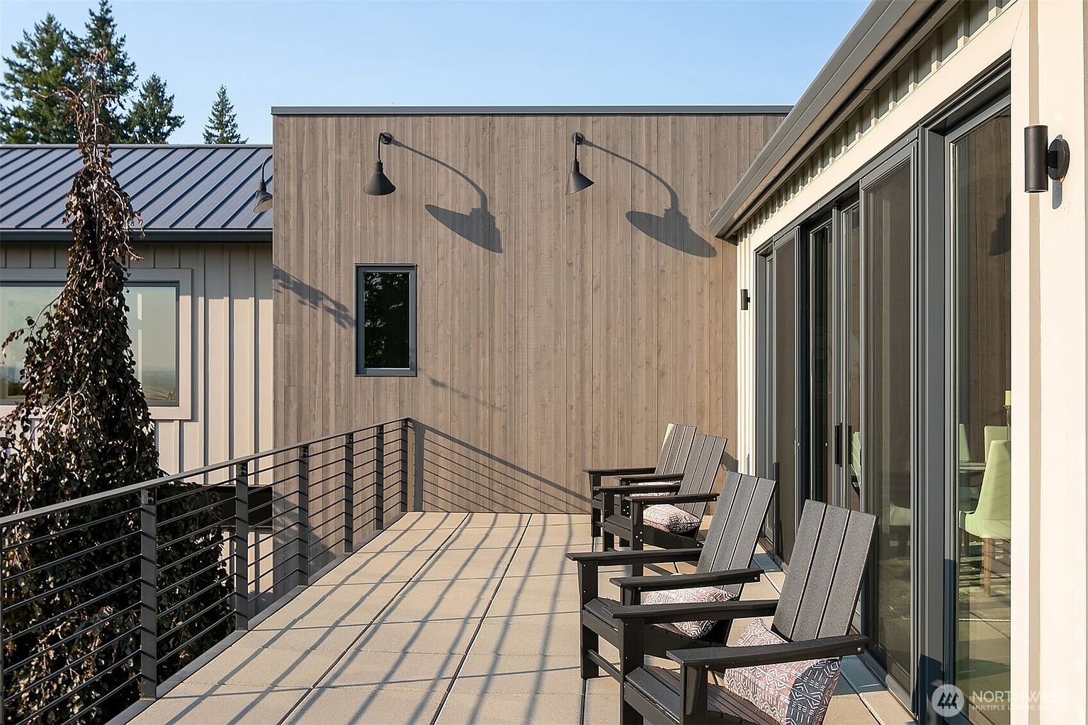 This modern, elevated deck features sleek, dark-toned Adirondack chairs arranged along a large sliding glass door, offering a seamless transition between indoor and outdoor living. The space is defined by clean lines, a minimalist metal railing, and warm-toned vertical wood siding, creating a sophisticated and contemporary atmosphere. The perspective captures the expansive deck surface and the architectural interplay of light and shadow on the exterior wall.