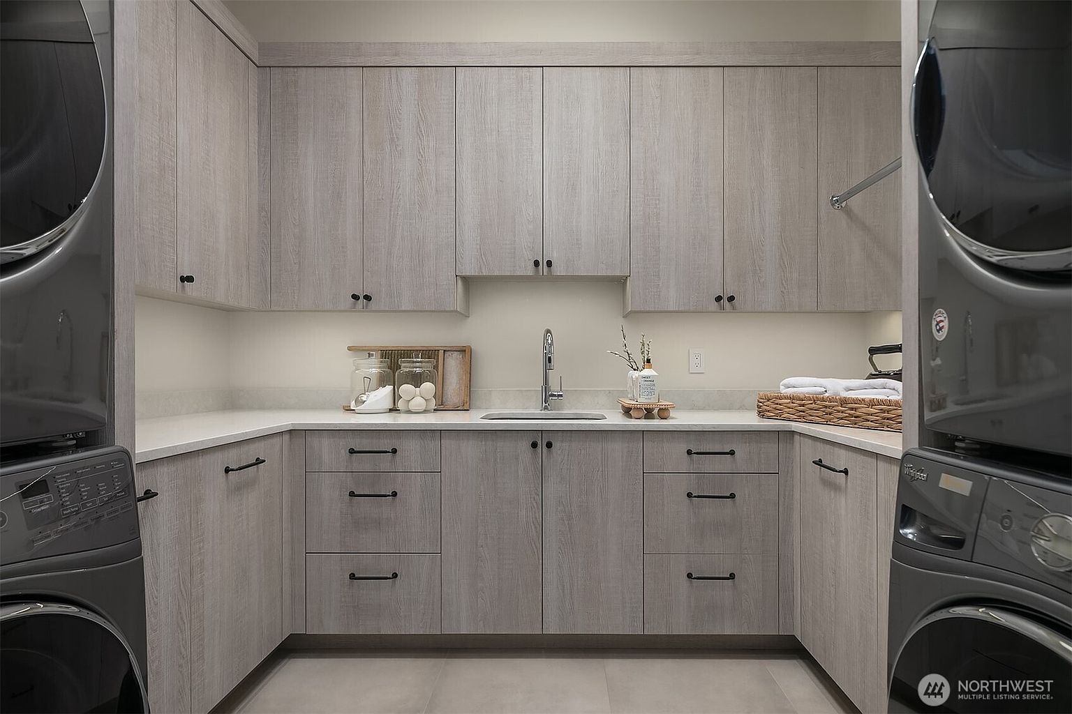 This modern, well-organized laundry room features light wood-grain cabinetry, sleek quartz countertops, and a stainless steel undermount sink. The space is framed by a stacked washer and dryer set on either side, creating a symmetrical and functional layout. The neutral color palette and clean lines provide a contemporary and polished aesthetic suitable for a high-end home.