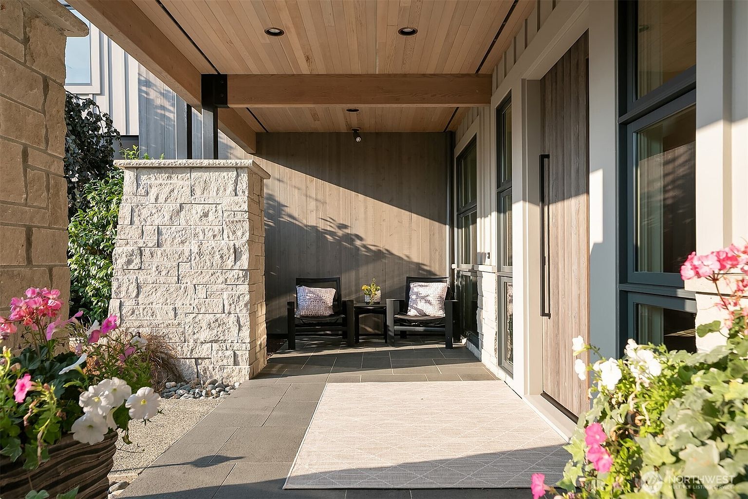 This inviting modern entryway features a covered porch with a warm wood-paneled ceiling and recessed lighting, creating a sophisticated and welcoming atmosphere. The space is accented by a textured stone pillar, a sleek wooden front door with a long vertical handle, and a pair of comfortable chairs with a small side table. Lush potted flowers in the foreground and a geometric-patterned outdoor rug add a touch of charm and comfort to this stylish home entrance.