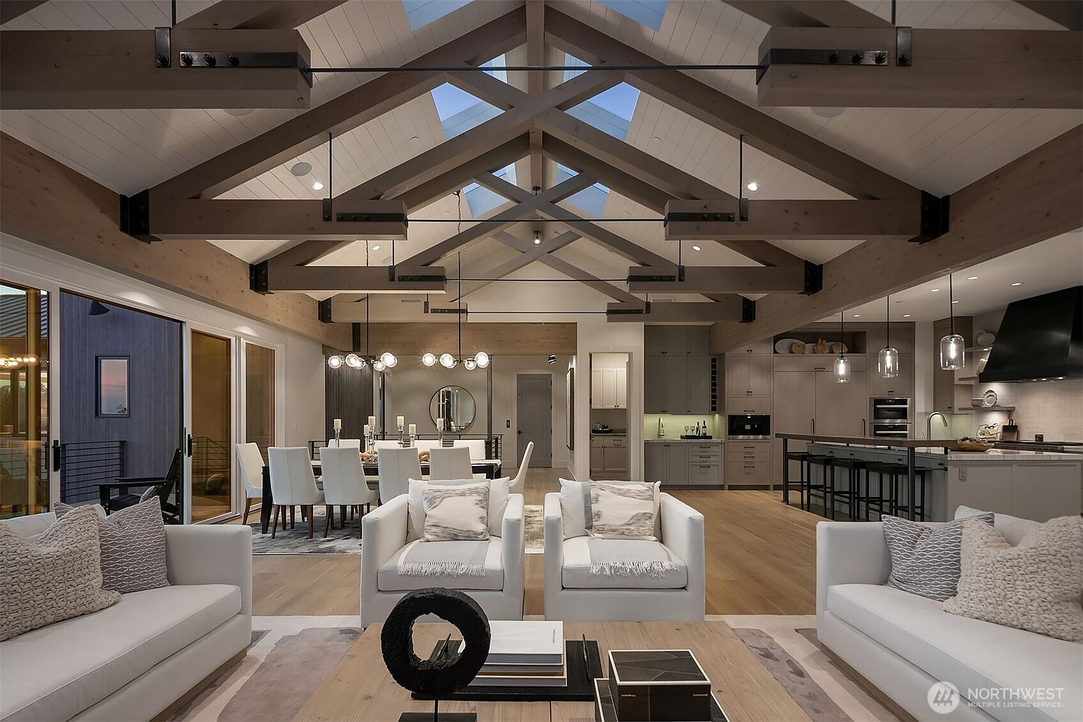 This expansive, open-concept living area features a dramatic vaulted ceiling with exposed timber beams and integrated skylights, creating a bright and airy atmosphere. The space seamlessly integrates a sophisticated living room with plush white seating, a formal dining area, and a modern kitchen, all anchored by light hardwood flooring. The design aesthetic is a refined blend of rustic warmth and contemporary luxury, perfect for high-end entertaining.