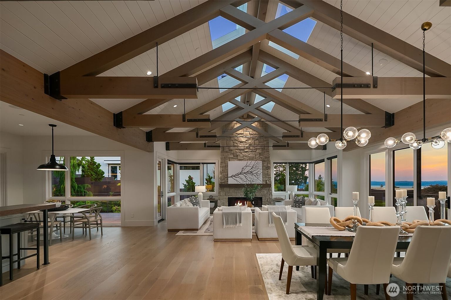 This expansive, open-concept living and dining area features a dramatic vaulted ceiling with exposed timber trusses and integrated skylights. The space is anchored by a grand stone fireplace and floor-to-ceiling windows that offer panoramic views of the water at sunset. The interior design blends rustic architectural elements with modern, sophisticated furnishings, creating an airy and luxurious atmosphere.