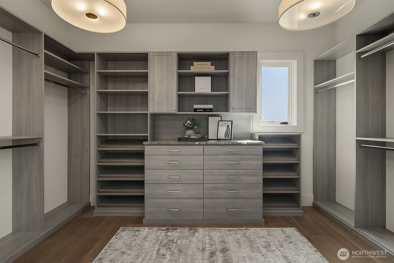 This sophisticated walk-in closet features custom grey wood cabinetry with a symmetrical layout, including ample shelving, hanging rods, and a central dresser with five drawers. The space is illuminated by two large, modern circular flush-mount ceiling lights and includes a small window that brings in natural light. A neutral-toned area rug sits on the hardwood floor, completing the organized and luxurious aesthetic.