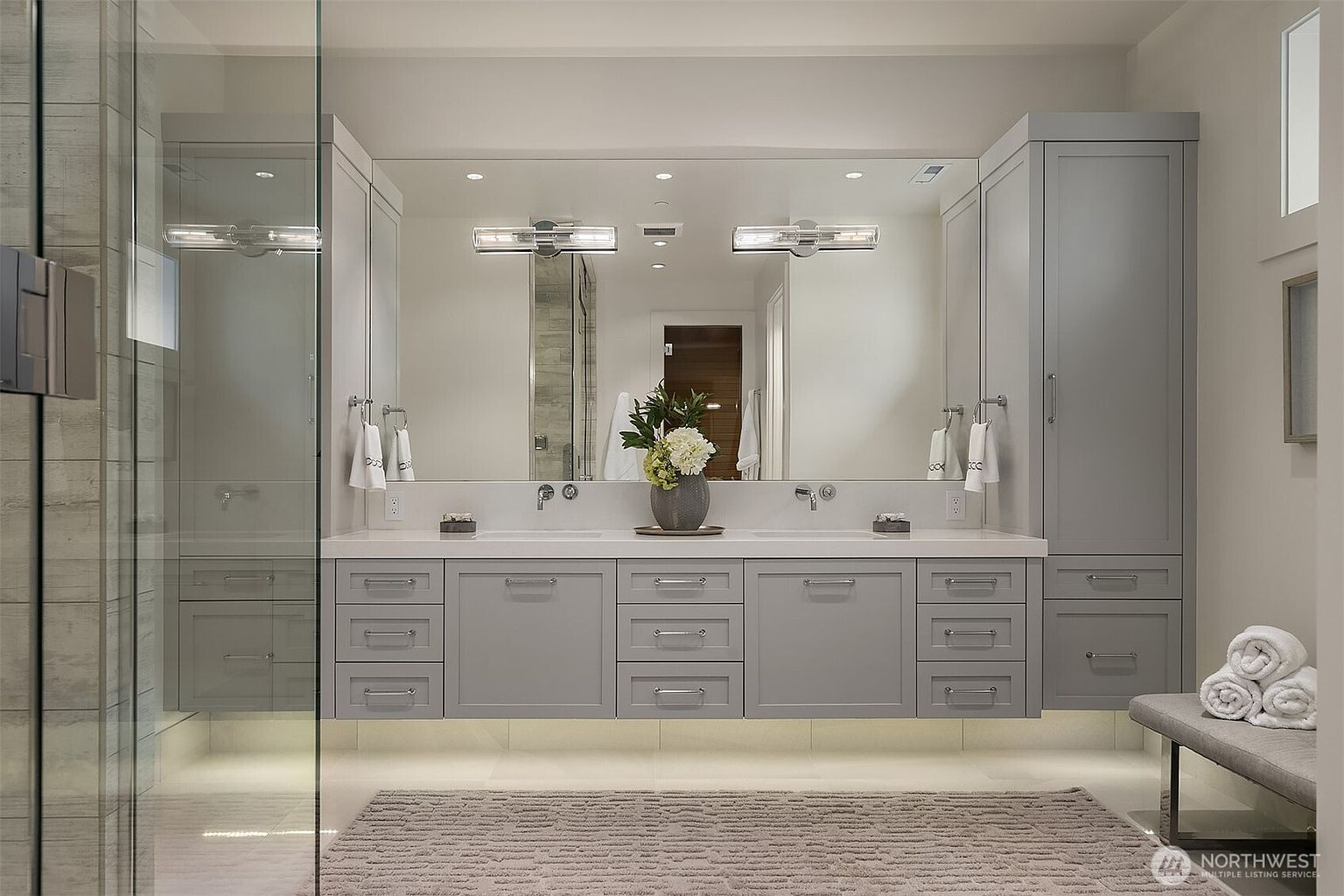 This elegant primary bathroom features a sleek, floating double vanity in a soft grey finish, topped with a clean white countertop and modern fixtures. A large, expansive mirror reflects the room, while under-cabinet lighting adds a warm, luxurious glow to the space. The room is completed by a glass-enclosed shower, a plush rug, and a bench with neatly folded towels, creating a serene and sophisticated spa-like atmosphere.