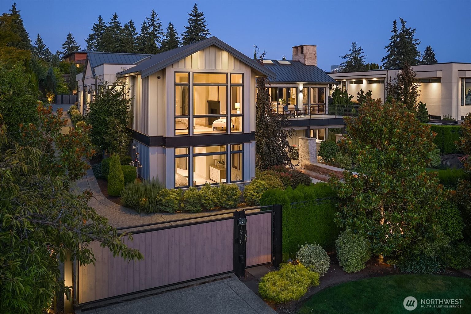 This striking contemporary residence is captured at dusk, showcasing a multi-level architectural design with expansive floor-to-ceiling windows that glow warmly from within. The property features a modern mix of vertical siding and stone accents, complemented by a meticulously landscaped garden and a private gated entrance. The elevated perspective highlights the home's sophisticated curb appeal and its seamless integration with the surrounding lush greenery.