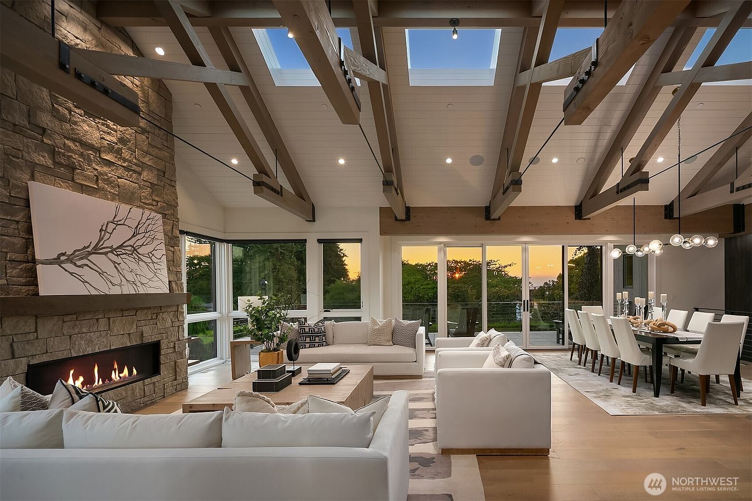 This expansive, open-concept living and dining area features a stunning vaulted ceiling with exposed timber beams and integrated skylights. A floor-to-ceiling stone fireplace serves as the focal point, complemented by modern white furnishings and large glass doors that offer a seamless transition to the outdoor space. The warm, natural light from the sunset creates an inviting and luxurious atmosphere, highlighting the high-end finishes and spacious layout.