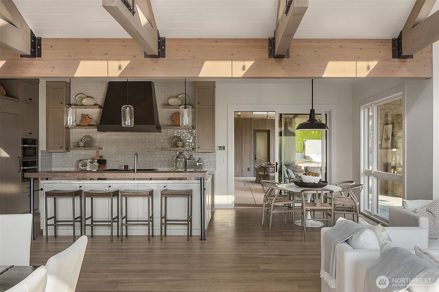 This bright, open-concept kitchen features a large island with bar seating, a striking dark range hood, and light wood ceiling beams that add a rustic, architectural touch. The space seamlessly transitions into a dining area and living room, with large sliding glass doors that invite natural light and provide a view of the outdoor patio. The design balances modern functionality with warm, organic textures, creating an inviting and airy atmosphere.