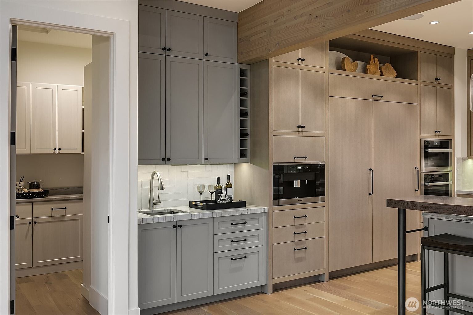 This sophisticated kitchen area features a high-end wet bar with grey cabinetry, marble-style countertops, and a built-in wine rack. Adjacent to the bar, a seamless wall of light-toned wood cabinetry integrates a built-in coffee machine and a large refrigerator, creating a clean, modern aesthetic. The perspective captures a functional and elegant transition space, highlighting the premium finishes and thoughtful design details.
