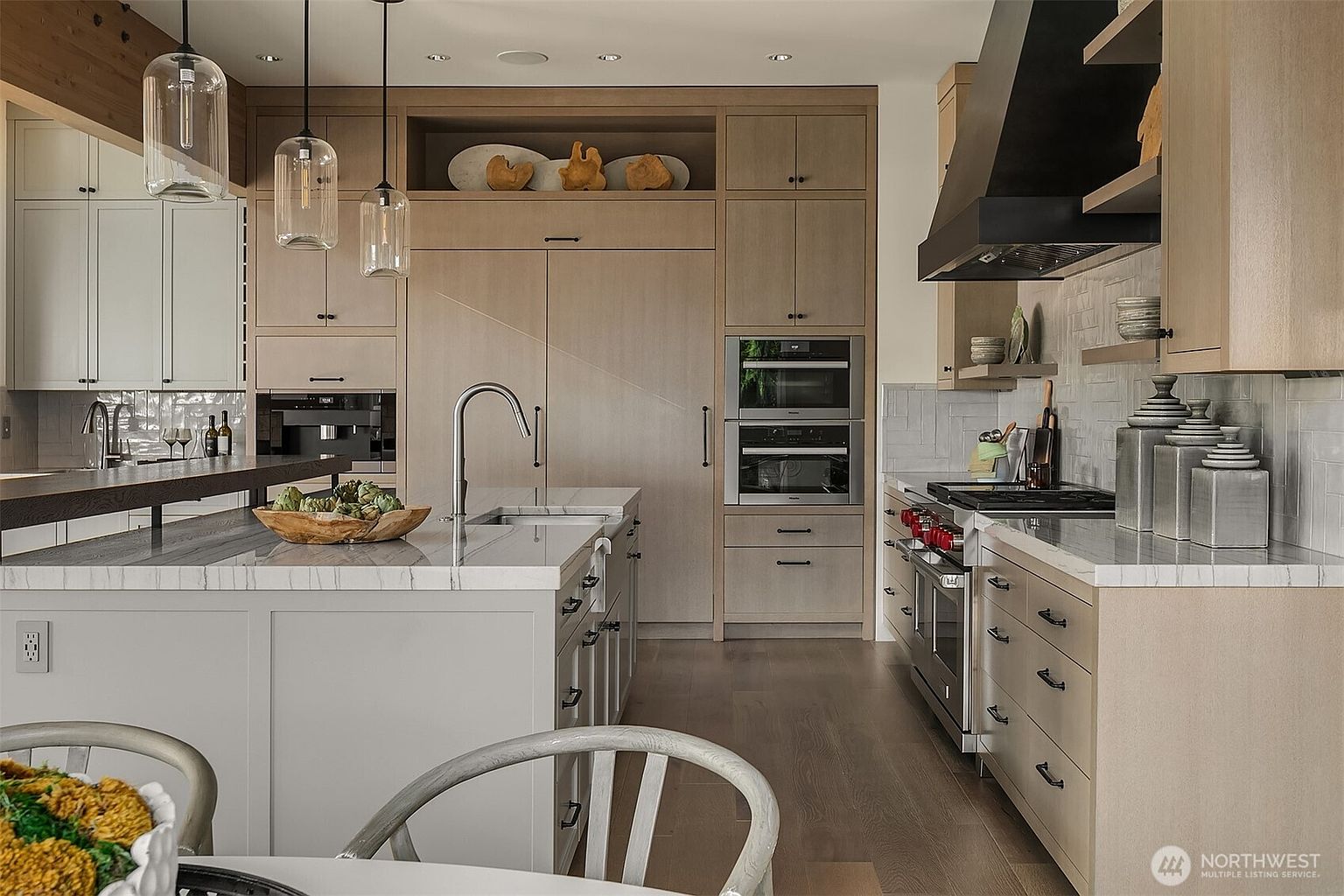 This sophisticated, modern kitchen features a seamless blend of light wood cabinetry and sleek, light-grey island cabinetry topped with white marble countertops. The space is equipped with high-end stainless steel appliances, including a built-in coffee machine, double wall ovens, and a professional-grade range with a custom hood. Pendant lighting hangs above the island, creating a warm and inviting atmosphere that highlights the clean lines and high-quality finishes throughout the room.