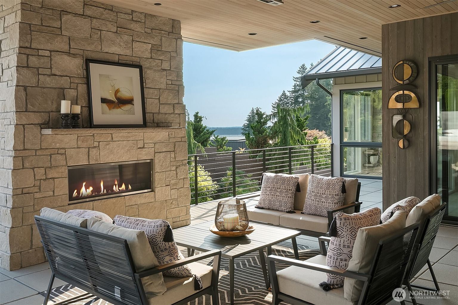 This luxurious covered outdoor living space features a modern linear fireplace built into a textured stone wall, creating a cozy focal point. The area is furnished with stylish, contemporary seating arranged around a central coffee table, all set against a backdrop of lush trees and a scenic water view. The space seamlessly blends indoor comfort with the beauty of the outdoors, perfect for relaxing or entertaining.