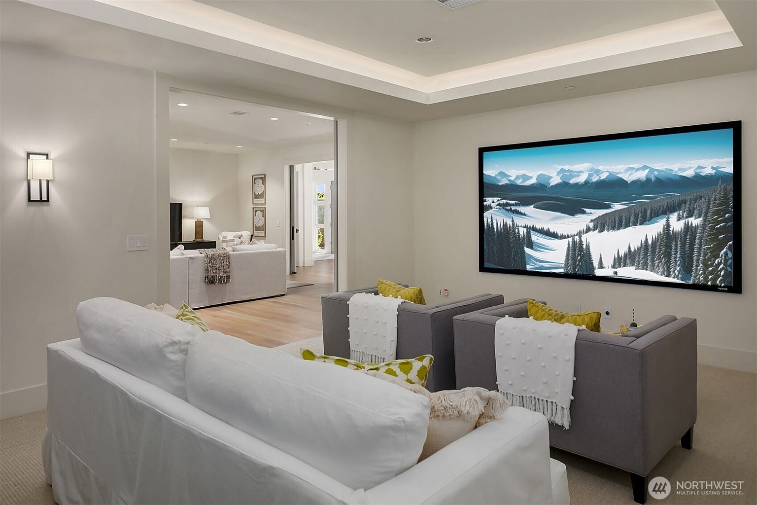 This sophisticated home theater room features a large wall-mounted screen displaying a scenic mountain landscape, complemented by plush gray armchairs and a comfortable white sofa. The room is designed with a modern, minimalist aesthetic, featuring recessed tray ceiling lighting and a seamless transition into an adjacent living area. The perspective is from the back of the room, highlighting the cozy, cinematic atmosphere perfect for entertainment.