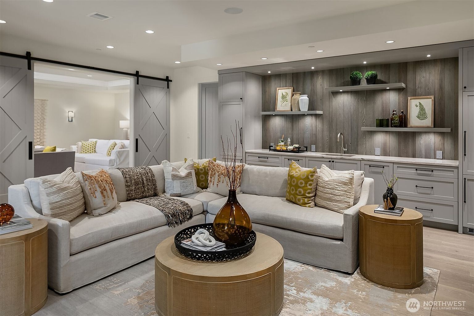 This sophisticated living room features a large, plush sectional sofa centered around a round wooden coffee table, creating a cozy and inviting atmosphere. The space is accented by a built-in wet bar with rustic wood paneling, open shelving, and elegant gray cabinetry, while sliding barn doors reveal a glimpse into an adjacent room. The neutral color palette, combined with warm lighting and textured decor, gives the room a refined, modern, and comfortable aesthetic.