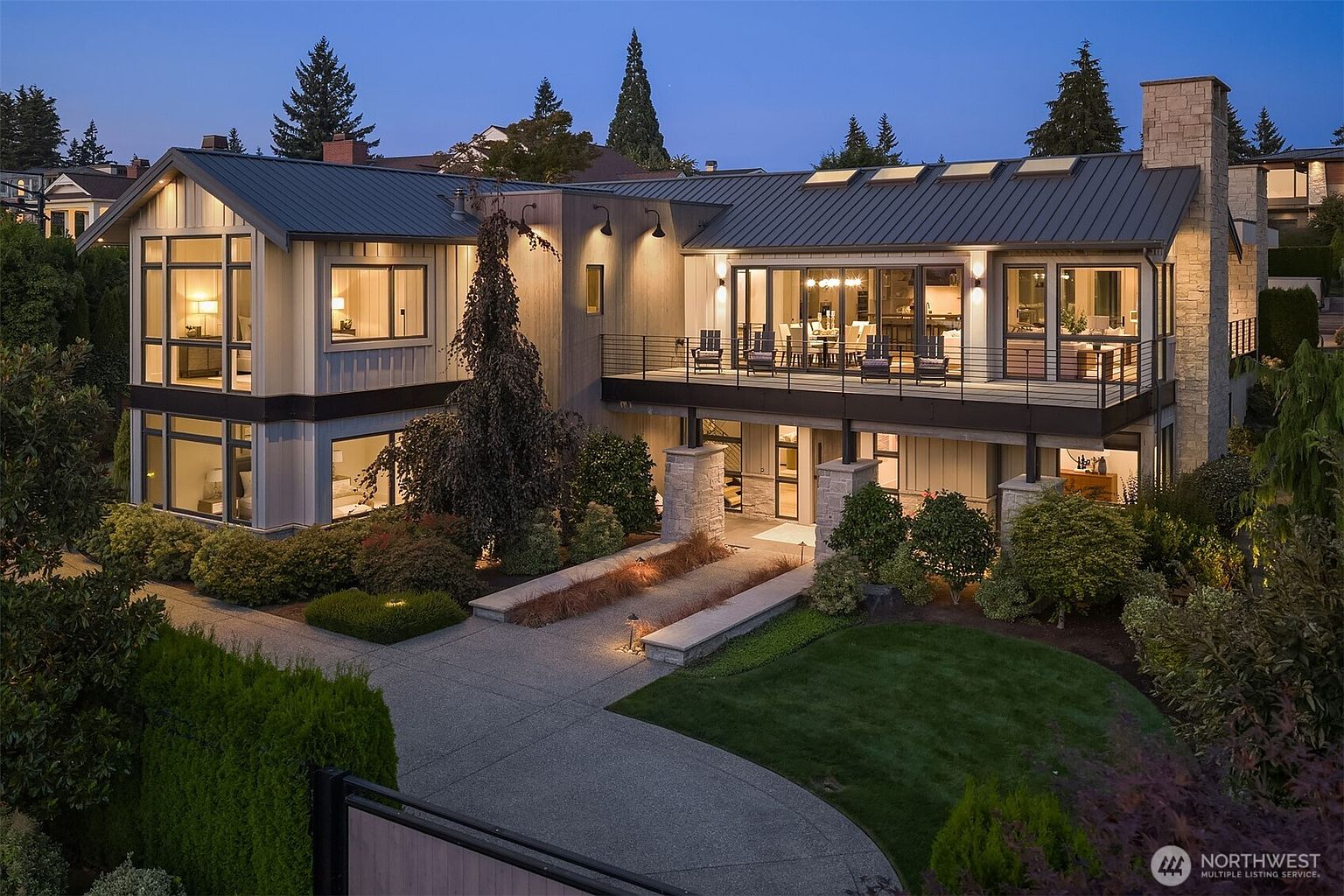 This stunning contemporary residence features a sophisticated blend of vertical wood siding, stone accents, and a sleek metal roof, captured during the golden hour. The home boasts a prominent elevated deck with glass railings, expansive floor-to-ceiling windows, and a beautifully landscaped front yard with a paved driveway. The warm exterior lighting creates an inviting and luxurious atmosphere, highlighting the architectural depth and modern design of the property.