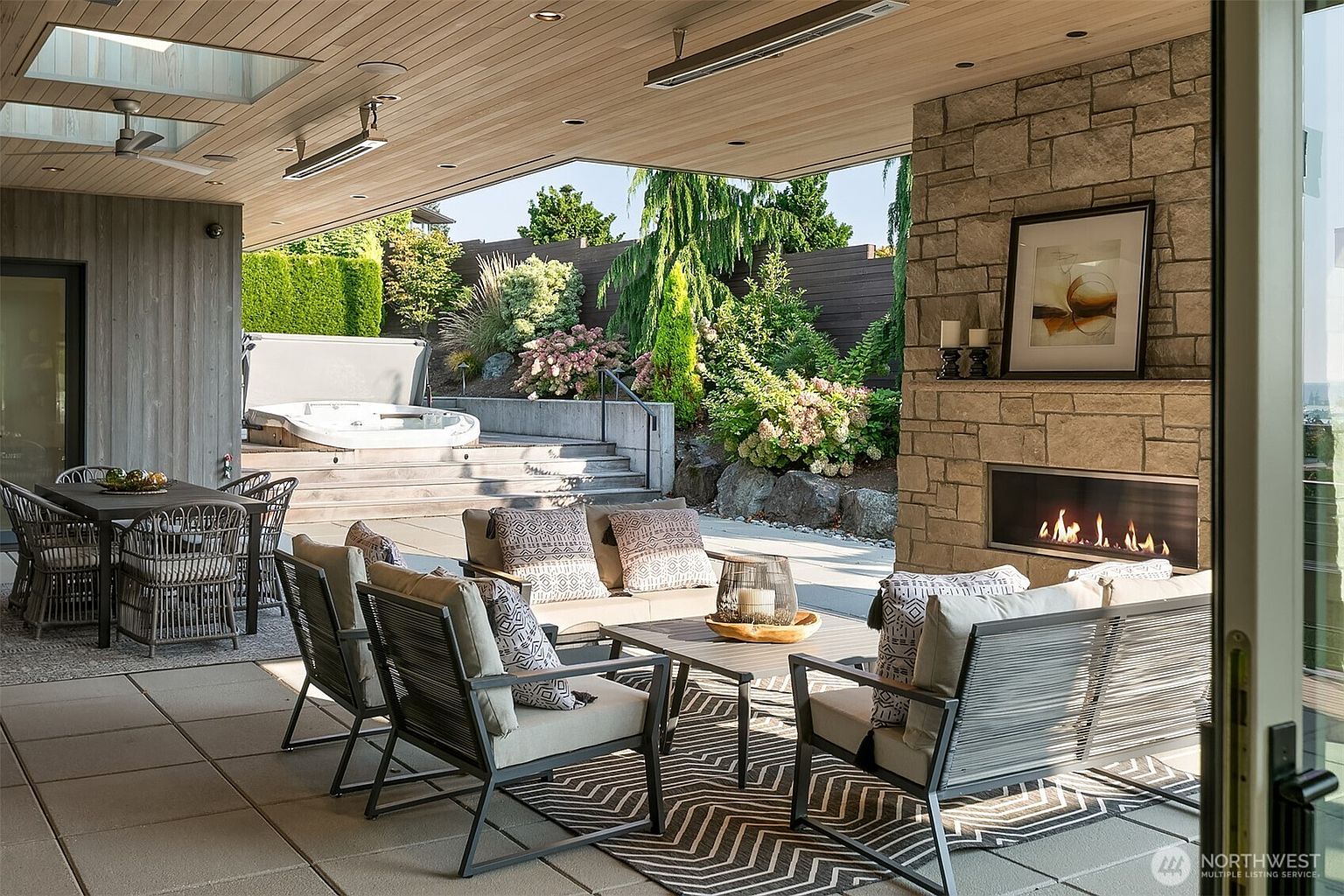 This luxurious covered outdoor living space features a modern stone fireplace, comfortable lounge seating, and a separate dining area, all set against a backdrop of lush, tiered landscaping. The space is seamlessly integrated with a built-in hot tub on the upper terrace, creating a sophisticated and inviting atmosphere for relaxation or entertaining. The architectural design emphasizes indoor-outdoor flow with high-end finishes and a serene, private garden setting.