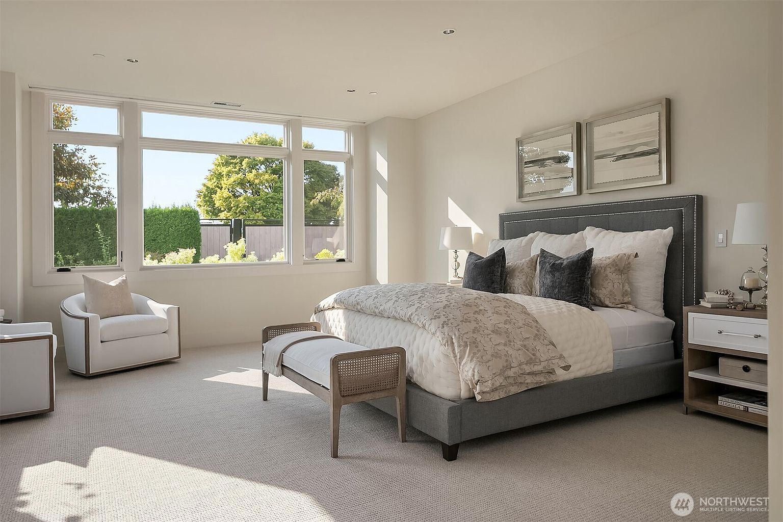 This elegant primary bedroom features a plush, neutral-toned color palette with a large, upholstered grey bed as the focal point. A comfortable armchair sits near a expansive window that floods the room with natural light, while a woven bench at the foot of the bed adds a touch of texture. The space is tastefully decorated with framed artwork and modern bedside lamps, creating a serene and sophisticated atmosphere.