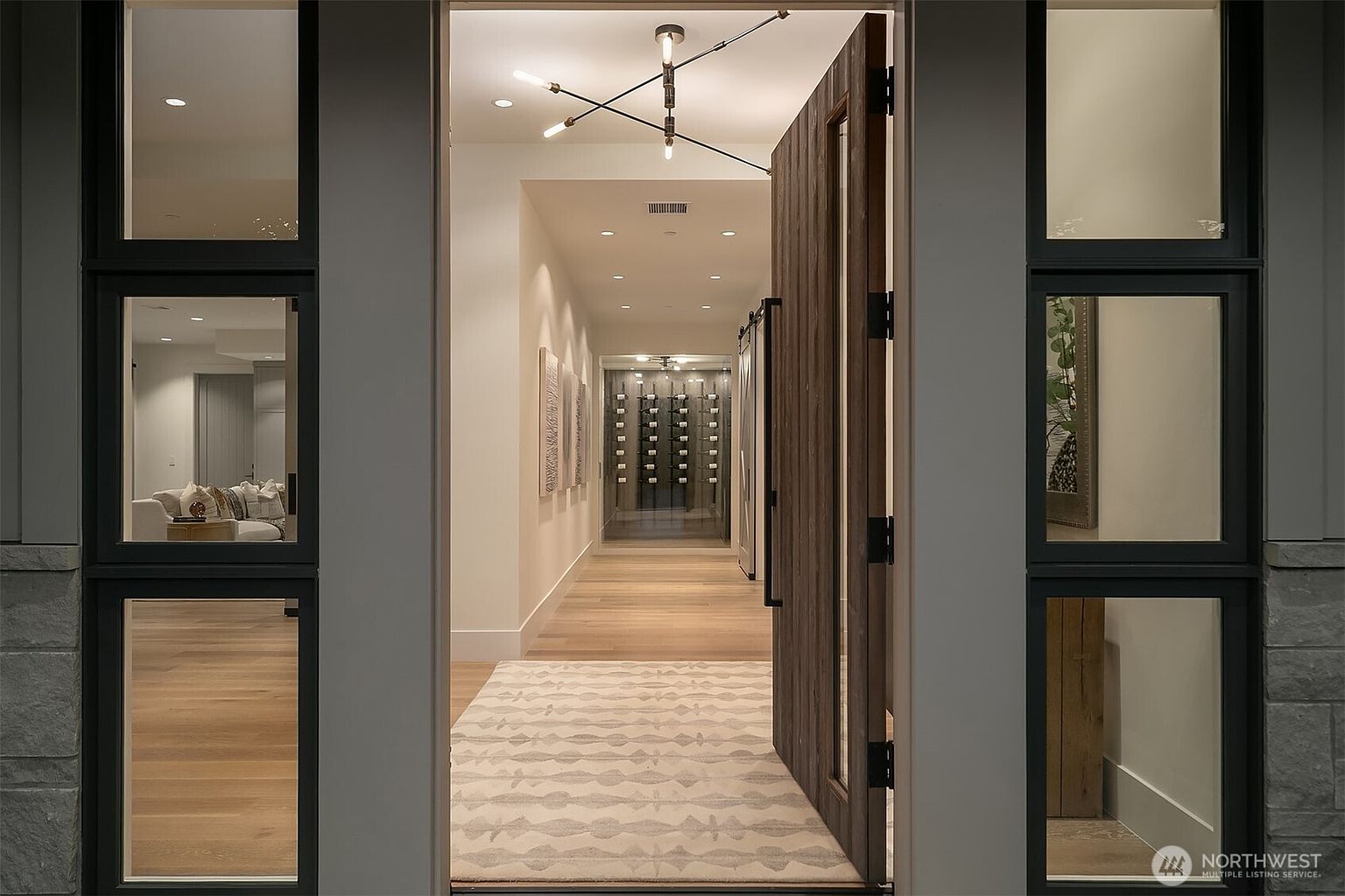 This perspective captures the grand entryway of a modern luxury home, looking through a large, pivot-style wooden front door into a bright, minimalist hallway. The interior features light hardwood flooring, a contemporary geometric chandelier, and a glass-enclosed wine display at the end of the corridor. The clean lines and sophisticated lighting create an inviting and high-end atmosphere, showcasing the seamless transition between the exterior and interior spaces.
