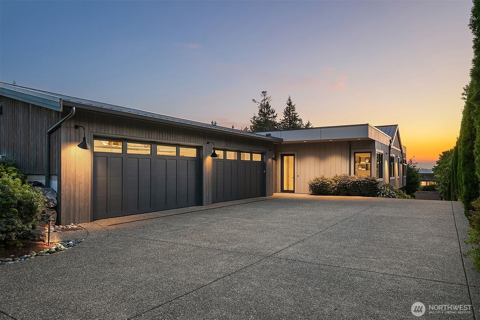 This modern home features a spacious three-car garage with dark, paneled doors and sleek exterior lighting, set against a backdrop of a vibrant sunset. The wide, aggregate concrete driveway leads up to the garage and a recessed entryway, showcasing a contemporary architectural style with clean lines and wood-paneled siding. The overall impression is one of sophisticated, high-end curb appeal in a serene, elevated setting.