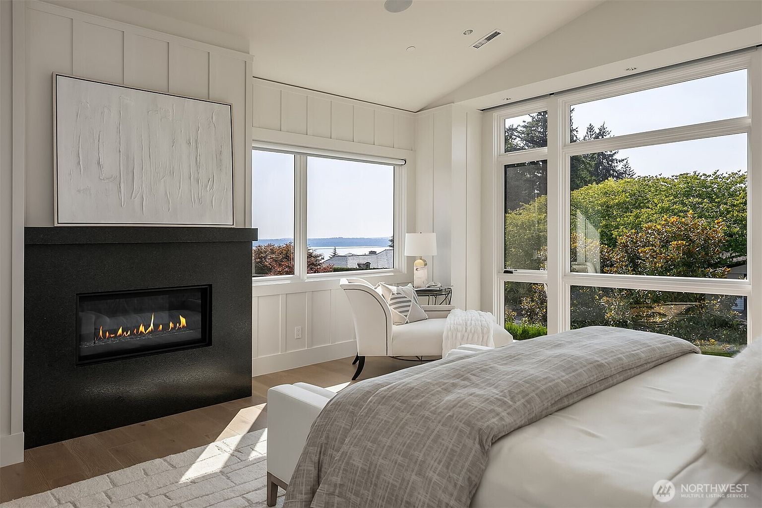 This elegant primary bedroom features a sophisticated, modern aesthetic with a striking black fireplace as a focal point, topped by a large abstract art piece. The room is bathed in natural light from expansive windows that offer a serene view of the water and lush greenery, complemented by a cozy reading nook with a white armchair. The space is finished with light hardwood floors and crisp white wall paneling, creating a tranquil and luxurious atmosphere.