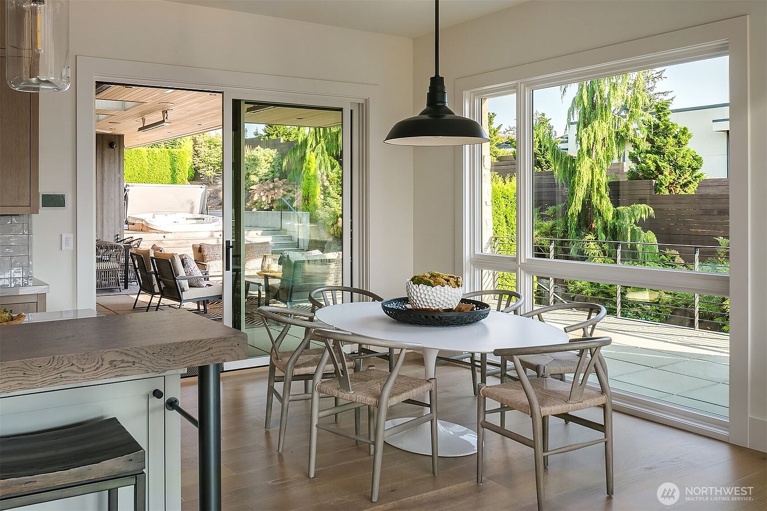 This bright and airy dining area features a modern white oval table surrounded by four wooden wishbone-style chairs, illuminated by a sleek black pendant light. Large sliding glass doors provide a seamless transition to an outdoor patio, creating an open-concept feel that connects the indoor living space with the lush greenery outside. The room is characterized by its clean lines, light wood flooring, and a neutral color palette that emphasizes natural light and a relaxed, contemporary atmosphere.