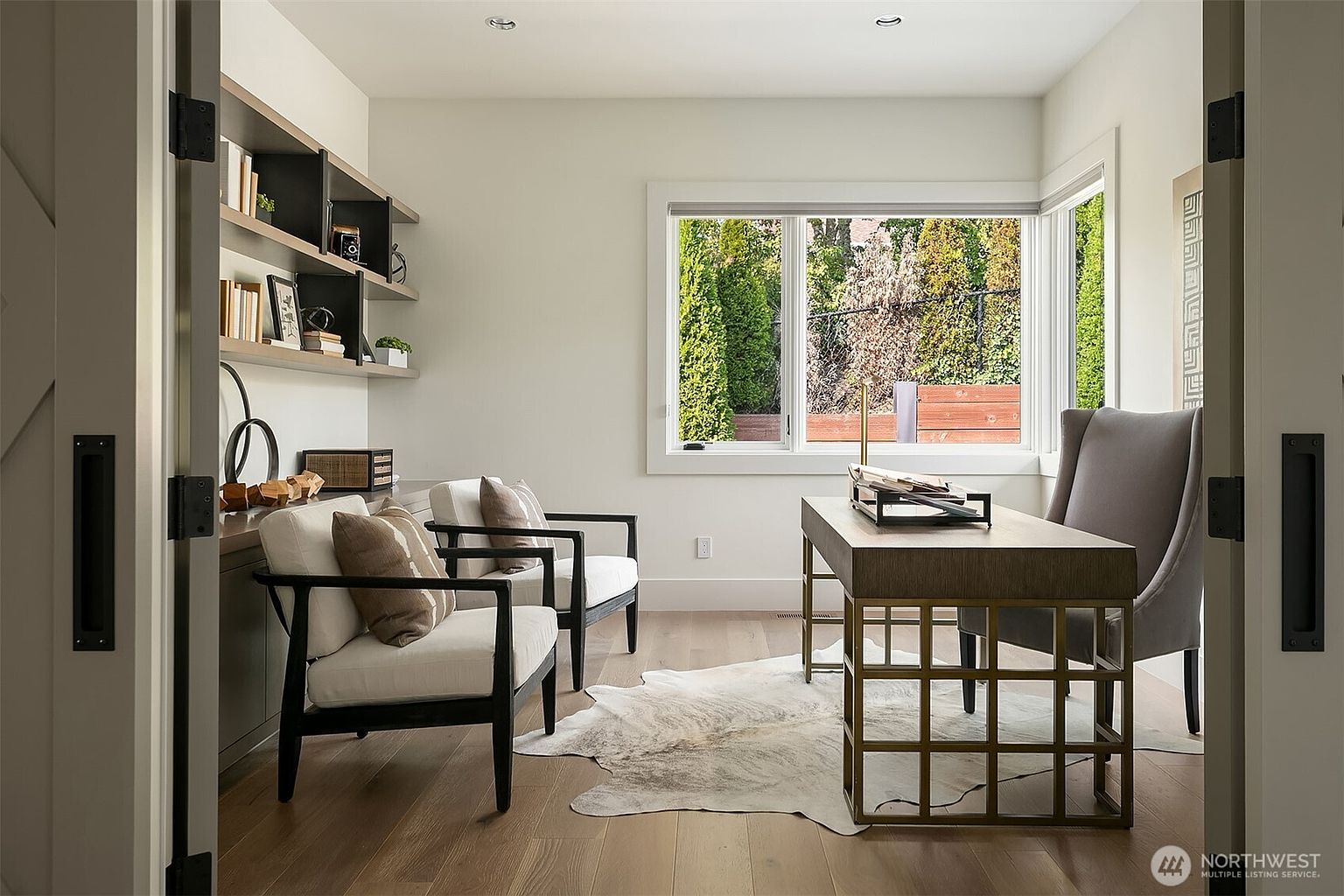 This sophisticated home office features a contemporary design with a sleek desk, two comfortable armchairs, and built-in shelving. The room is bathed in natural light from a large window overlooking a lush, green backyard, creating a serene and productive atmosphere. A stylish cowhide rug anchors the space, while the neutral color palette and clean lines provide a modern, professional aesthetic.