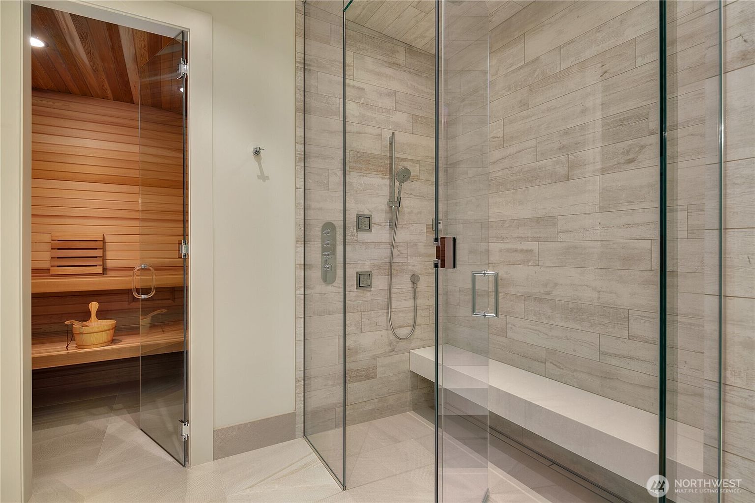 This luxurious bathroom features a modern glass-enclosed walk-in shower with light-toned, wood-look tile walls and a built-in bench. Adjacent to the shower is a dedicated sauna room with warm cedar wood paneling and a wooden bucket, creating a high-end spa-like atmosphere. The perspective captures both the functional shower area and the relaxing sauna, highlighting the premium amenities of the space.