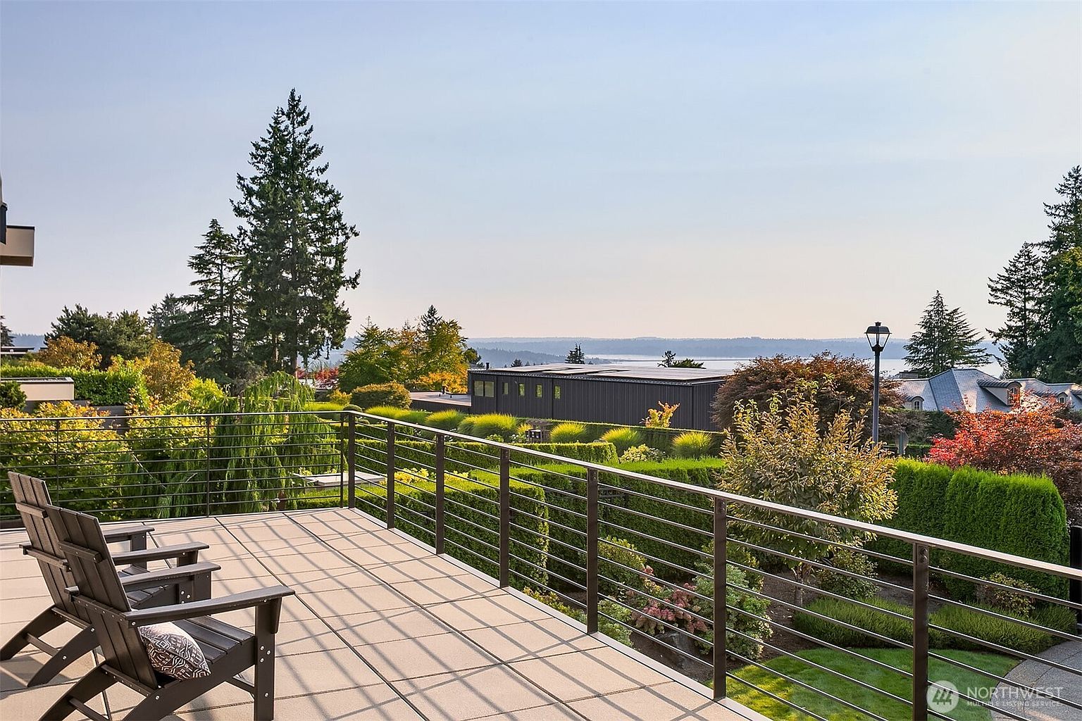 This elevated deck offers a serene, panoramic view of the surrounding landscape, including manicured hedges, lush trees, and a distant body of water. The space is furnished with two dark wooden Adirondack chairs, providing a perfect spot for relaxation while overlooking the well-maintained garden below. The modern metal railing and clean, paved surface create a sophisticated and inviting outdoor living area.