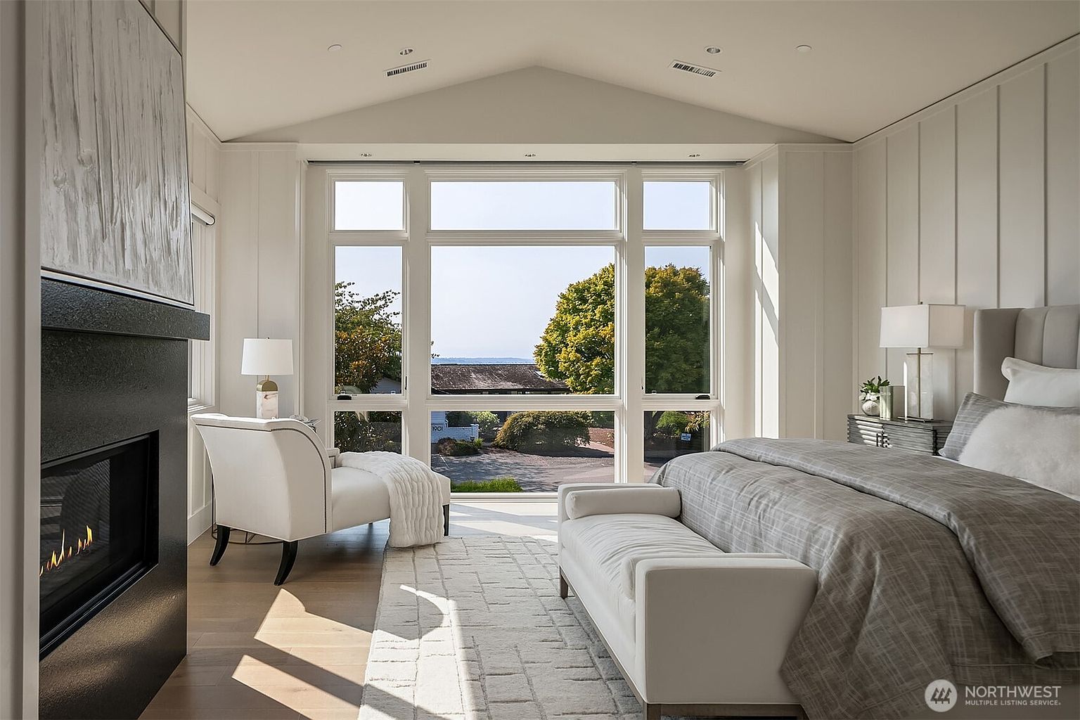 This elegant primary bedroom features a vaulted ceiling, a cozy fireplace, and a large window wall that frames a scenic outdoor view. The room is furnished with a plush bed, a stylish bench, and a comfortable armchair, all set against a neutral, sophisticated color palette. The bright, airy atmosphere and high-end finishes create a serene and luxurious retreat.