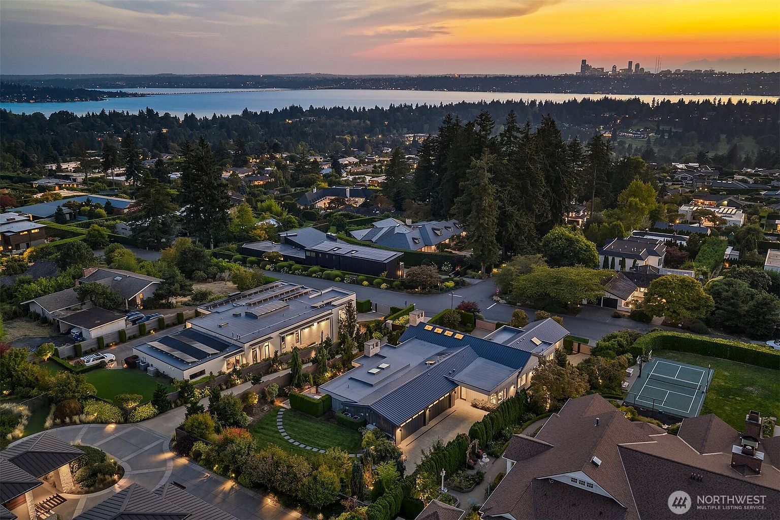 This stunning aerial view captures a luxurious residential neighborhood at sunset, showcasing modern homes with sleek metal roofs and solar panels. The perspective highlights the expansive property layouts, manicured landscaping, and a private tennis court, all set against a breathtaking backdrop of a lake and a distant city skyline. The warm, golden hour lighting enhances the sophisticated architectural design and the serene, upscale atmosphere of the community.
