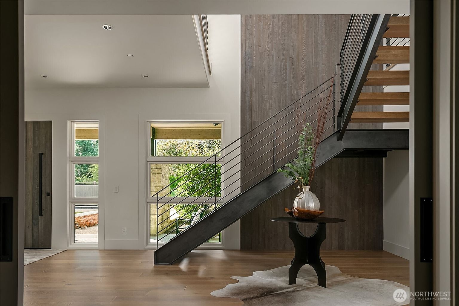 This striking entryway features a dramatic floating staircase with a sleek metal frame and warm wood treads, set against a textured, dark wood-paneled accent wall. The space is brightened by large, multi-pane windows that offer a view of the outdoors, while a minimalist round table with a decorative vase sits atop a cowhide rug. The overall aesthetic is a sophisticated blend of modern industrial design and natural warmth.