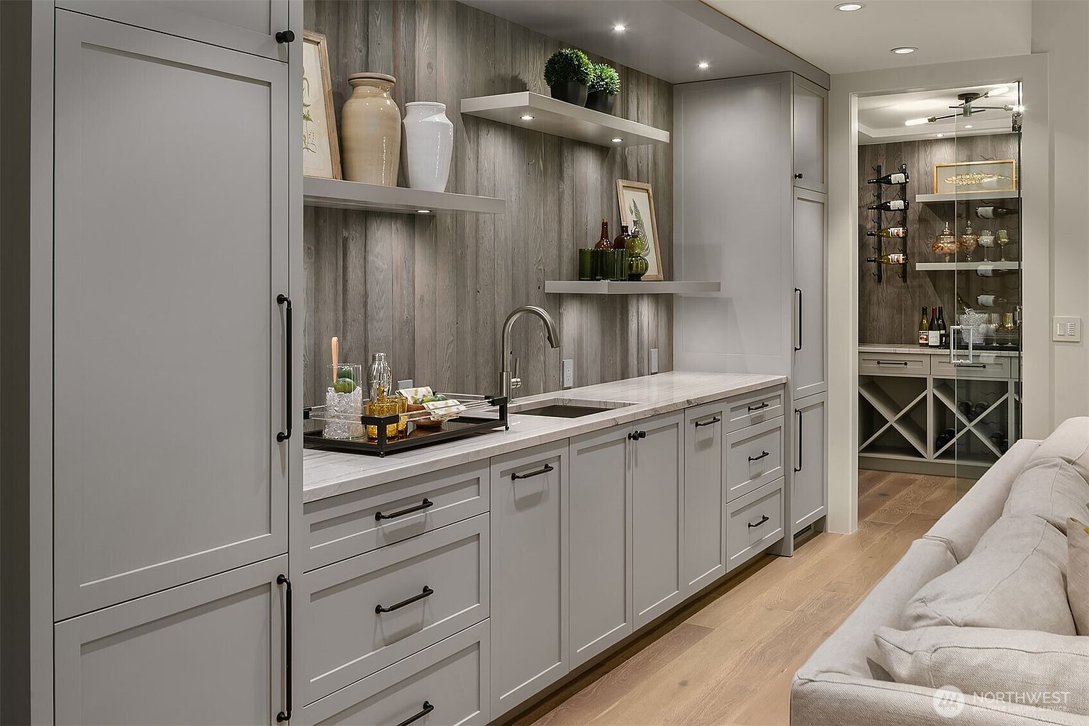 This sophisticated wet bar area features sleek grey cabinetry, a marble countertop, and a rustic wood-paneled backsplash that adds warmth to the modern design. The space is accented by floating shelves, recessed lighting, and a glass-enclosed wine storage room visible in the background. The composition offers a clear, eye-level perspective of a high-end, functional entertaining space.