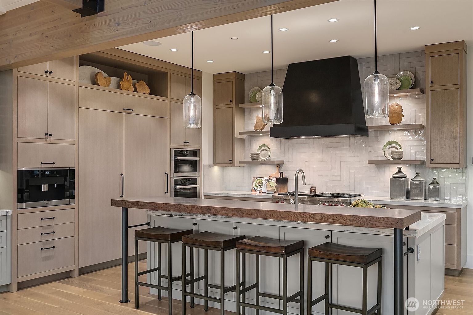 This sophisticated, modern-rustic kitchen features light wood cabinetry, a large island with a thick wood countertop, and a striking black range hood. The space is accented by a herringbone-patterned tile backsplash, open shelving, and elegant glass pendant lights, creating a warm and inviting atmosphere. The perspective is a straight-on, eye-level shot that highlights the high-end finishes and functional layout of the culinary space.