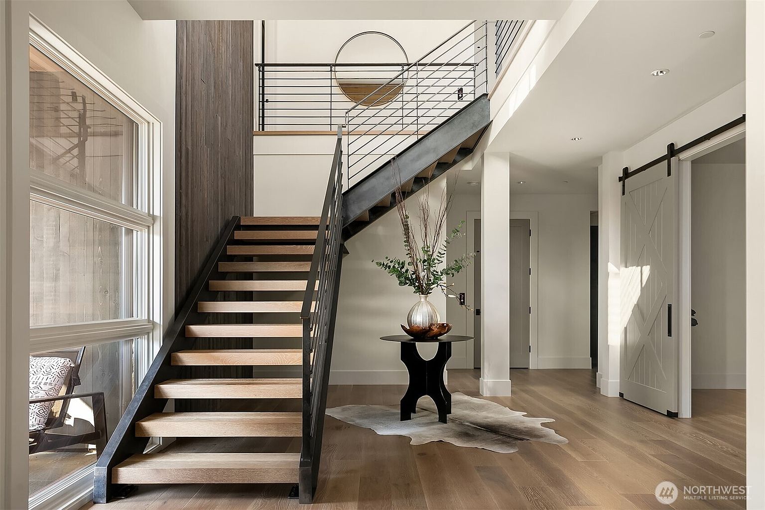 This striking entryway features a modern floating staircase with light wood treads and a sleek black metal railing, serving as the focal point of the home. A circular mirror hangs on the landing above, while a sculptural pedestal table with a floral arrangement sits on a cowhide rug below, creating a sophisticated and welcoming atmosphere. The space is bright and airy, characterized by clean lines, high ceilings, and a sliding barn door that adds a touch of rustic-modern charm.