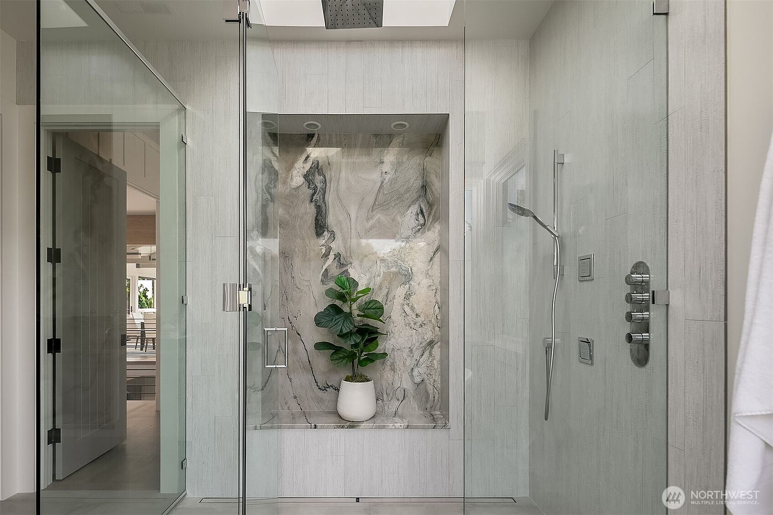 This luxurious walk-in shower features a sophisticated design with light-toned, textured wall tiles and a striking marble-accented recessed niche housing a decorative potted plant. The space is enclosed by seamless glass panels and includes a modern rainfall showerhead, a handheld sprayer, and multiple wall-mounted control valves. The overall aesthetic is clean, contemporary, and spa-like, emphasizing high-end finishes and meticulous craftsmanship.