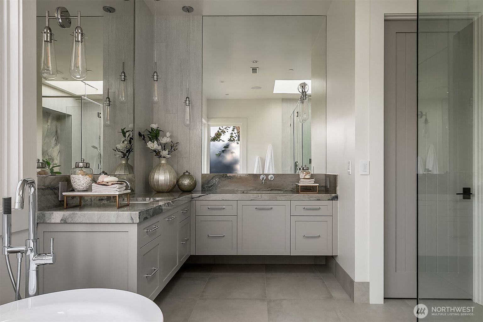 This elegant primary bathroom features a sleek, L-shaped floating vanity with light gray cabinetry and a striking marble countertop. A large, frameless mirror spans the wall, reflecting the modern pendant lighting and a glimpse of a skylight, while a glass-enclosed shower area adds to the open, airy feel. The space is finished with neutral, large-format floor tiles, creating a sophisticated and tranquil atmosphere.
