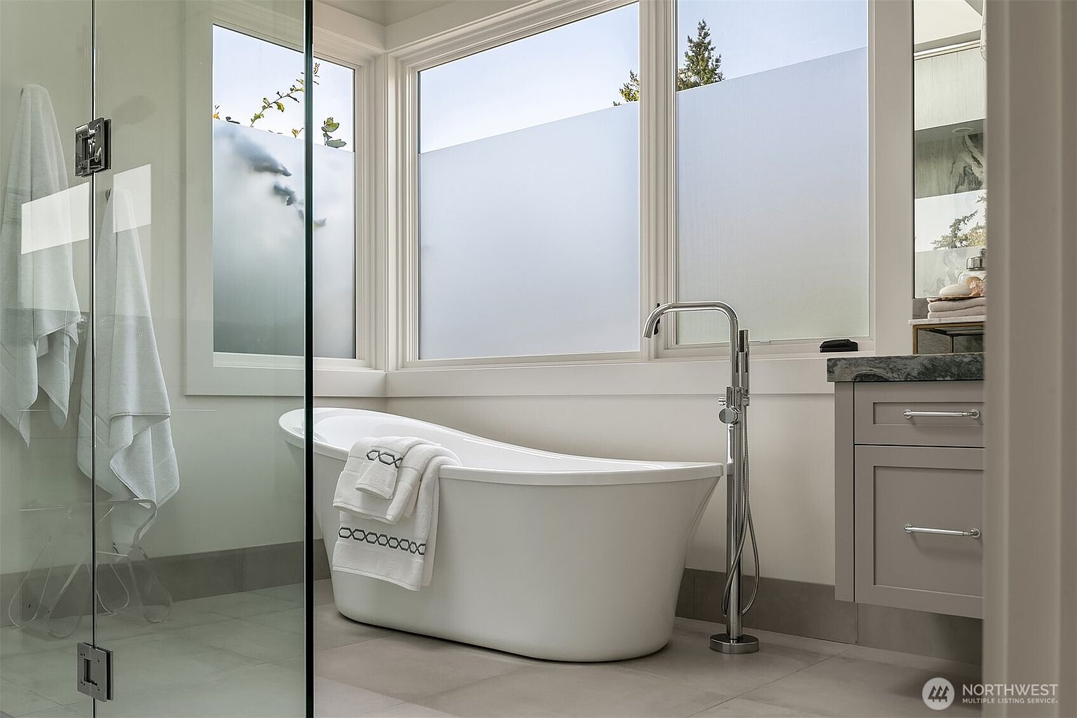 This elegant primary bathroom features a sleek, white freestanding soaking tub positioned beneath large windows with frosted glass for privacy. To the left, a glass-enclosed shower is visible, while a modern vanity with a stone countertop sits to the right, complemented by a tall, polished chrome floor-mounted tub filler. The space exudes a clean, contemporary aesthetic with neutral tones and high-end finishes, creating a serene and spa-like atmosphere.