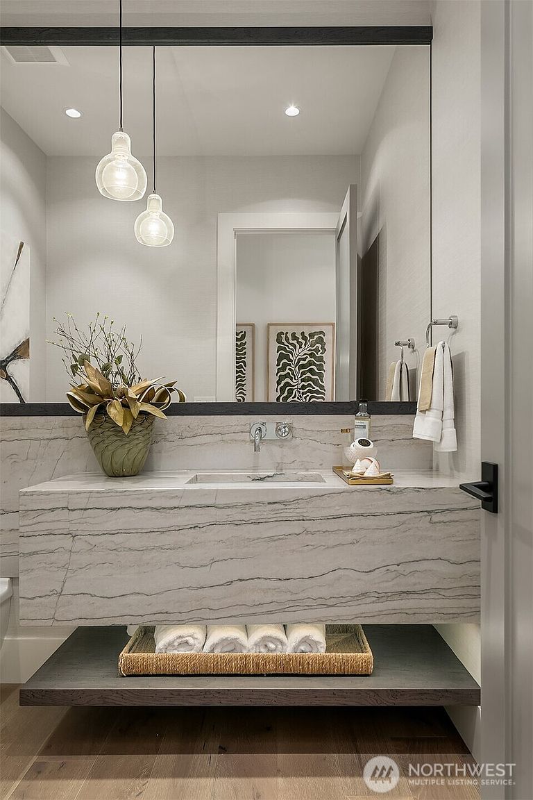 This elegant powder room features a striking floating vanity crafted from veined marble, complemented by a dark wood lower shelf holding neatly rolled towels. A large, full-width mirror reflects the space, accented by two modern glass pendant lights and a decorative plant arrangement. The minimalist design is enhanced by a wall-mounted faucet and a clean, sophisticated color palette.