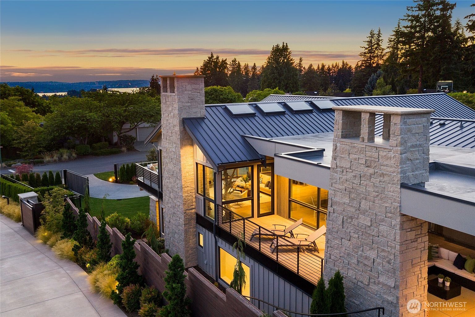 This high-angle aerial shot captures a sophisticated modern home at dusk, highlighting its striking architectural blend of stone chimneys, dark metal roofing, and expansive glass walls. The perspective emphasizes the seamless transition between the indoor living spaces and the elevated deck, which overlooks a beautifully landscaped yard and a distant water view. The warm interior lighting creates an inviting contrast against the cool tones of the twilight sky, showcasing a luxurious and contemporary aesthetic.