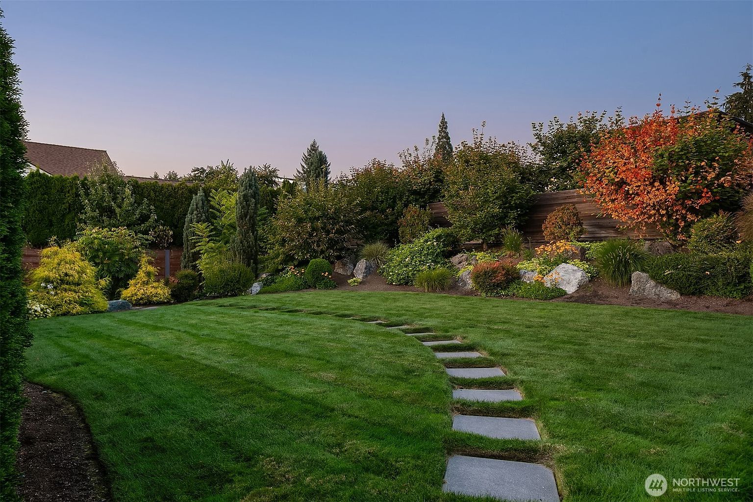 This beautifully landscaped backyard features a lush, manicured green lawn with a curved stone paver path leading toward a vibrant garden bed. The garden is densely planted with a variety of shrubs, trees, and colorful foliage, set against a backdrop of a wooden privacy fence. The perspective is a low-angle, eye-level shot that emphasizes the depth and tranquility of the outdoor space during the golden hour.