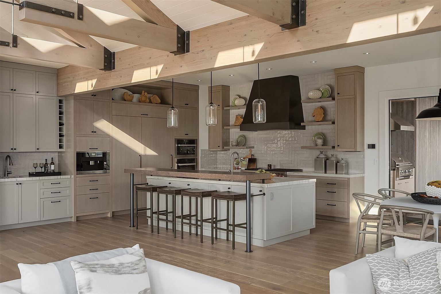 This sophisticated, open-concept kitchen features a blend of light wood cabinetry and sleek, modern design elements, anchored by a large central island with bar seating. The space is highlighted by exposed timber ceiling beams, a striking black range hood, and elegant pendant lighting, creating a warm yet contemporary atmosphere. The perspective is a wide, eye-level shot that captures the seamless flow between the kitchen, dining area, and living space, emphasizing the home's airy and high-end aesthetic.
