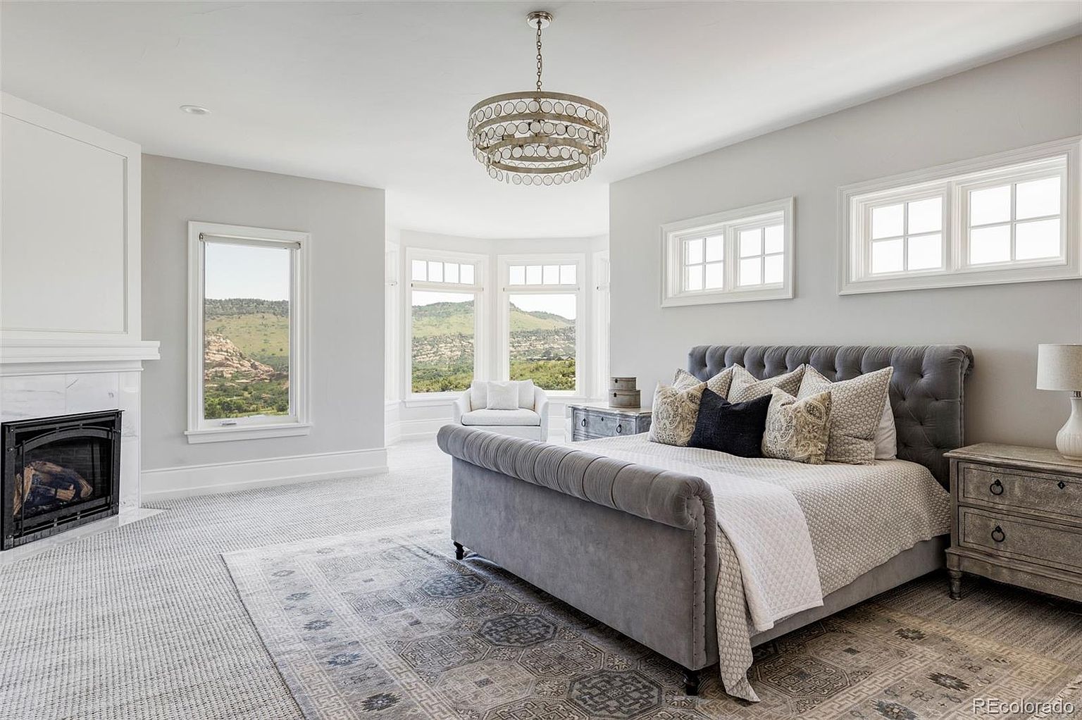 This is a luxurious primary bedroom featuring a plush, tufted gray bed with decorative pillows and a white throw. The room is bathed in natural light from multiple windows offering scenic views, and a stylish chandelier hangs from the ceiling. A fireplace adds a cozy touch, and the overall aesthetic is elegant and serene.