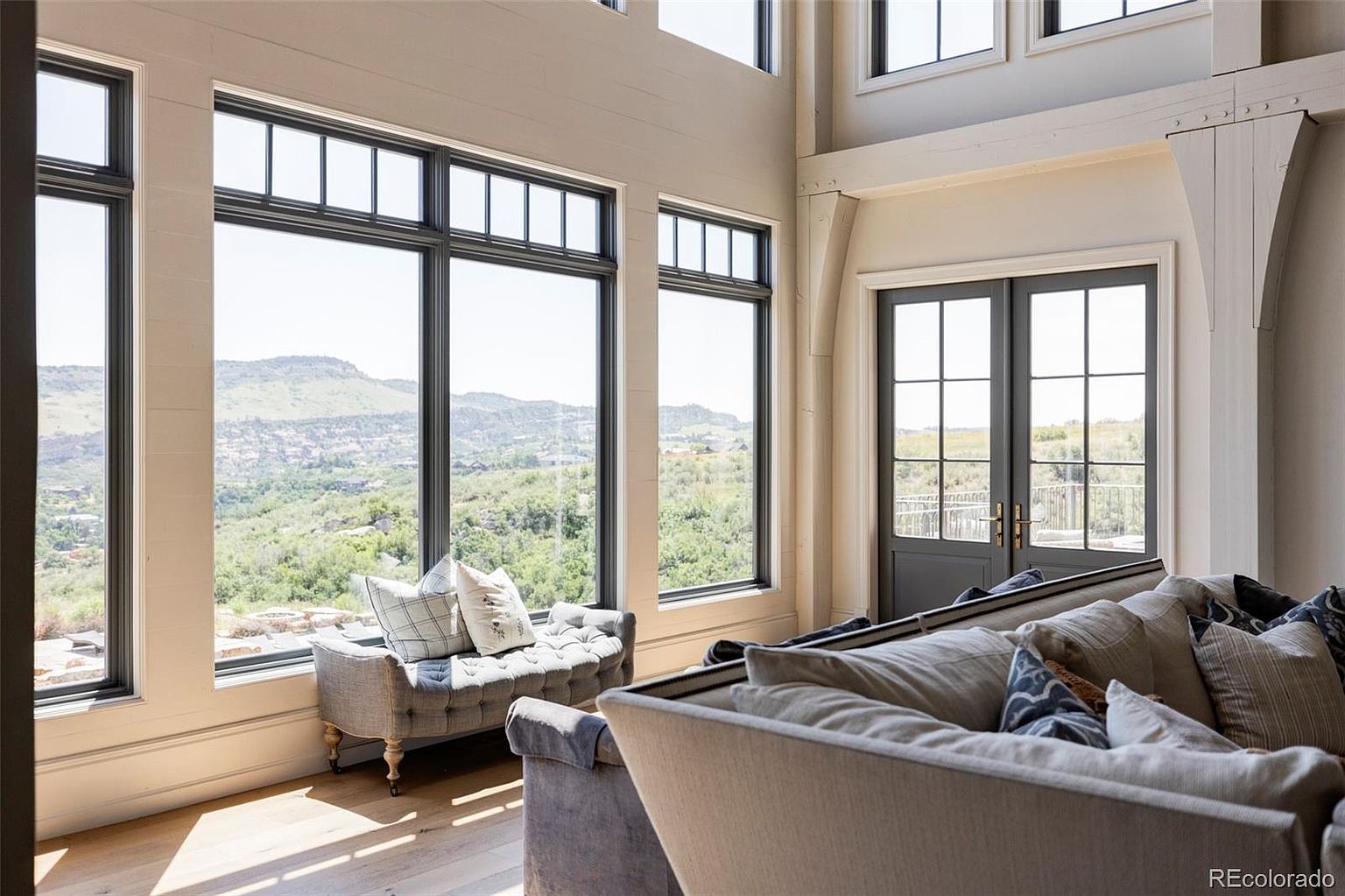 This is an interior shot of a living room featuring large windows with black frames that offer a scenic view of rolling hills and greenery. A tufted bench sits beneath the windows, and a comfortable sofa with numerous pillows is in the foreground. The room has a bright and airy feel, with light-colored walls and hardwood floors, creating a welcoming and luxurious atmosphere.