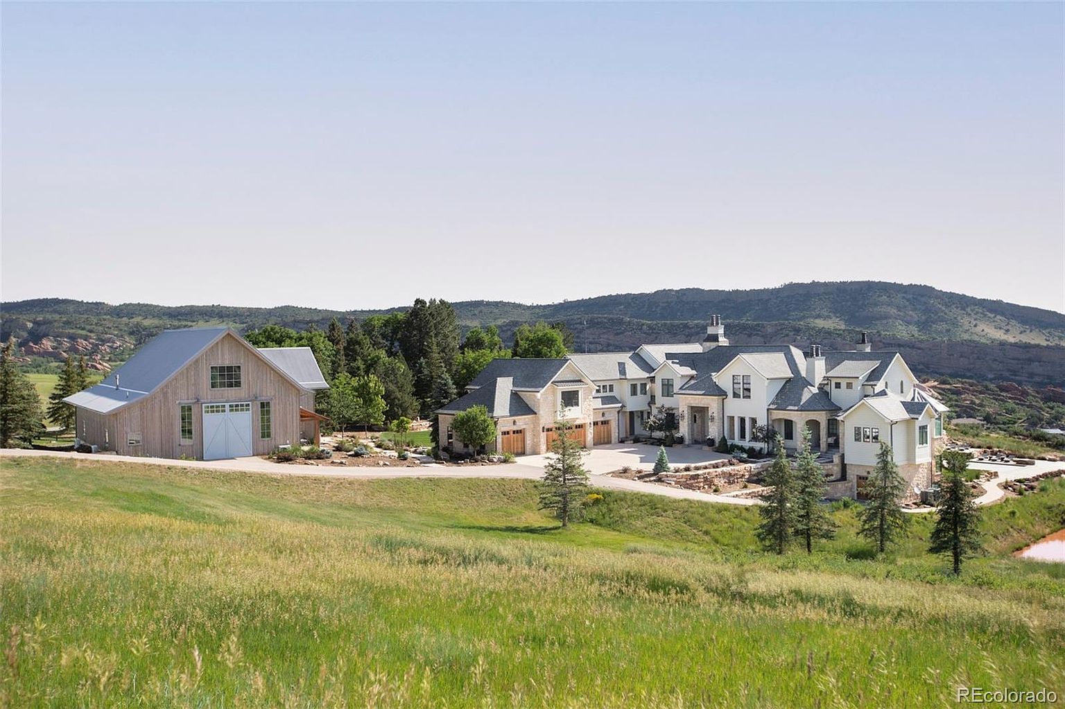 This image showcases the front view of a luxurious estate, featuring a large, modern farmhouse-style home with white exteriors and a stone accent garage. A separate barn-like structure complements the main house, set against a backdrop of rolling hills and a clear blue sky. The property includes a well-manicured lawn and landscaping, creating a serene and upscale ambiance.
