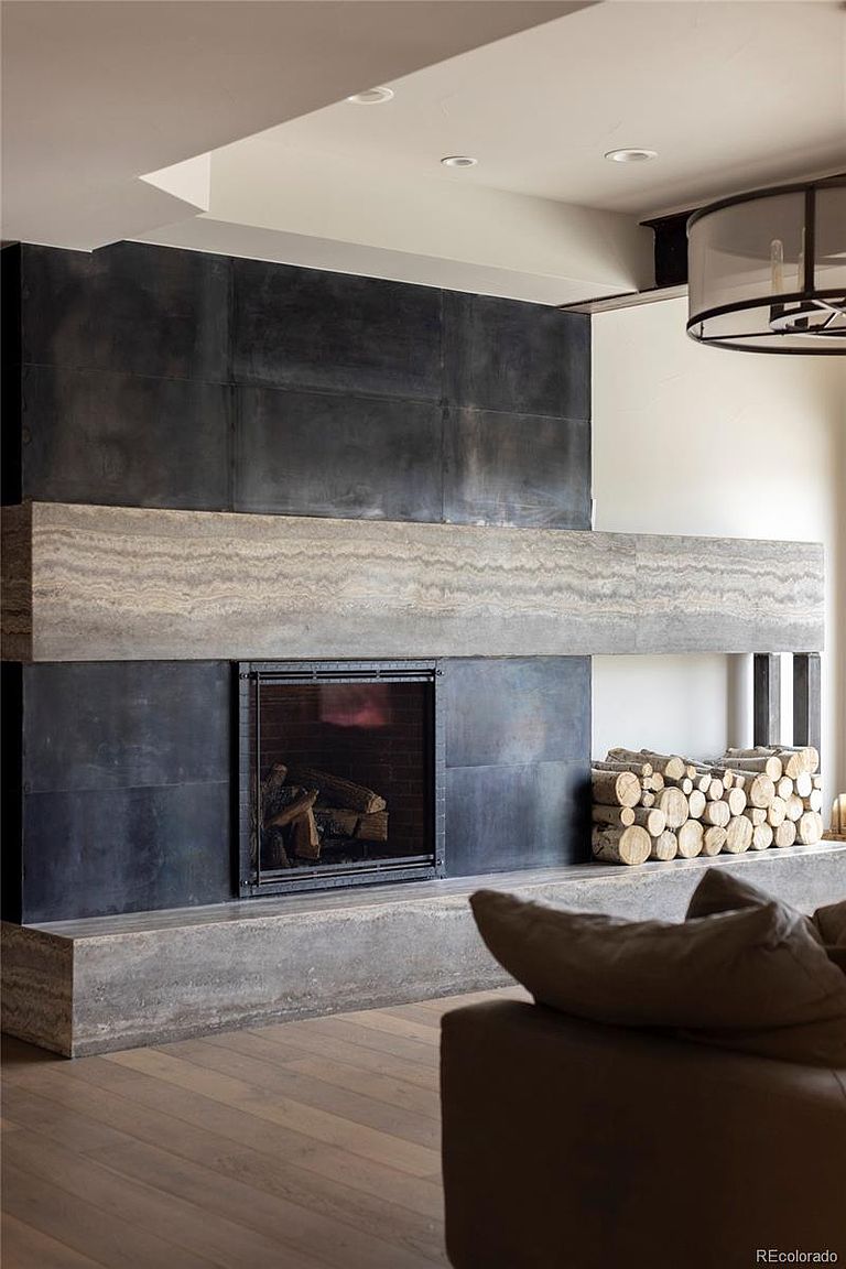 This interior shot showcases a modern living room with a striking fireplace as the focal point. The fireplace features a dark metal surround, a light-colored stone mantel, and a visible firebox with logs. A stack of firewood is neatly arranged to the side, and a portion of a sofa with pillows is visible in the foreground, suggesting a cozy and inviting atmosphere.