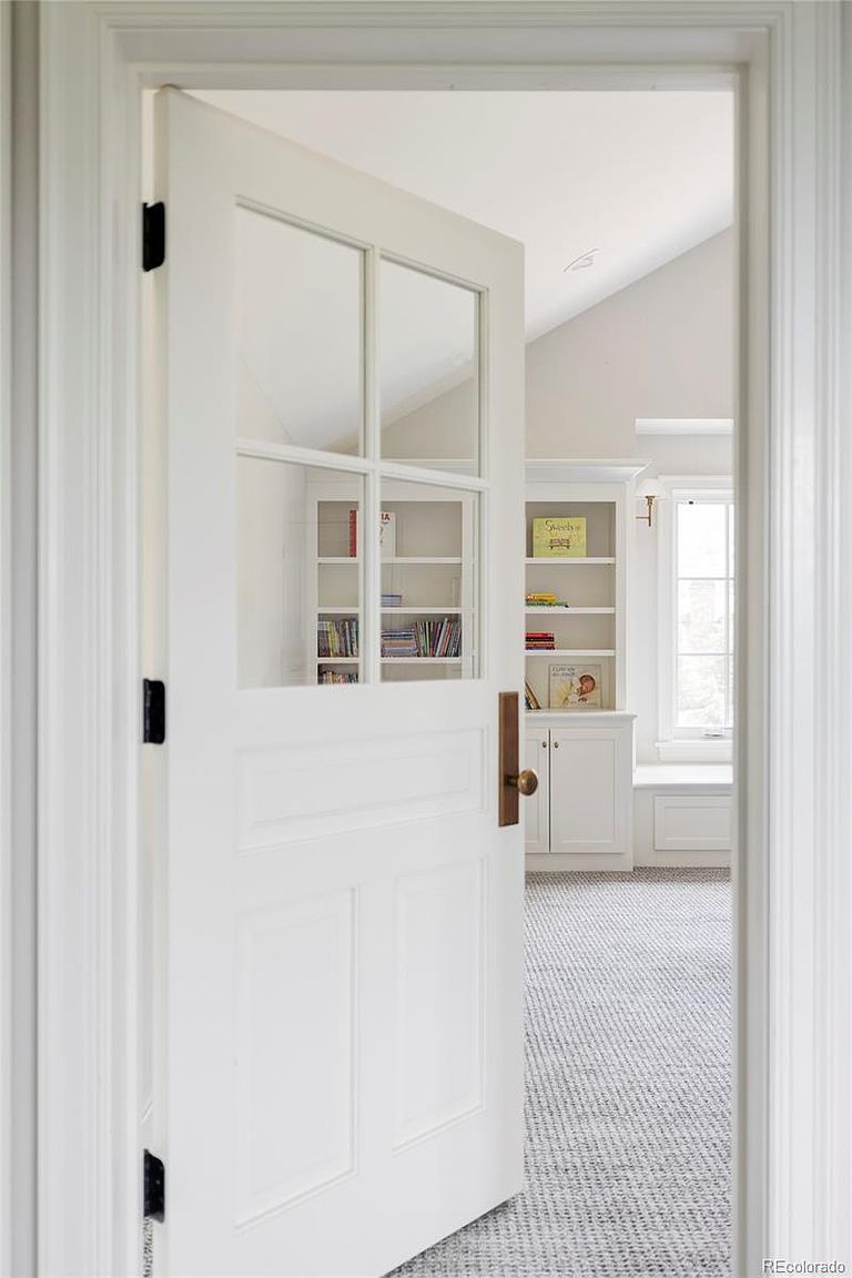 The image showcases an office or study, viewed through a partially open white paneled door. Inside, built-in white bookshelves are visible, filled with books and decorative items. The room is carpeted in a neutral tone, and natural light streams in from a window, creating a bright and inviting workspace.