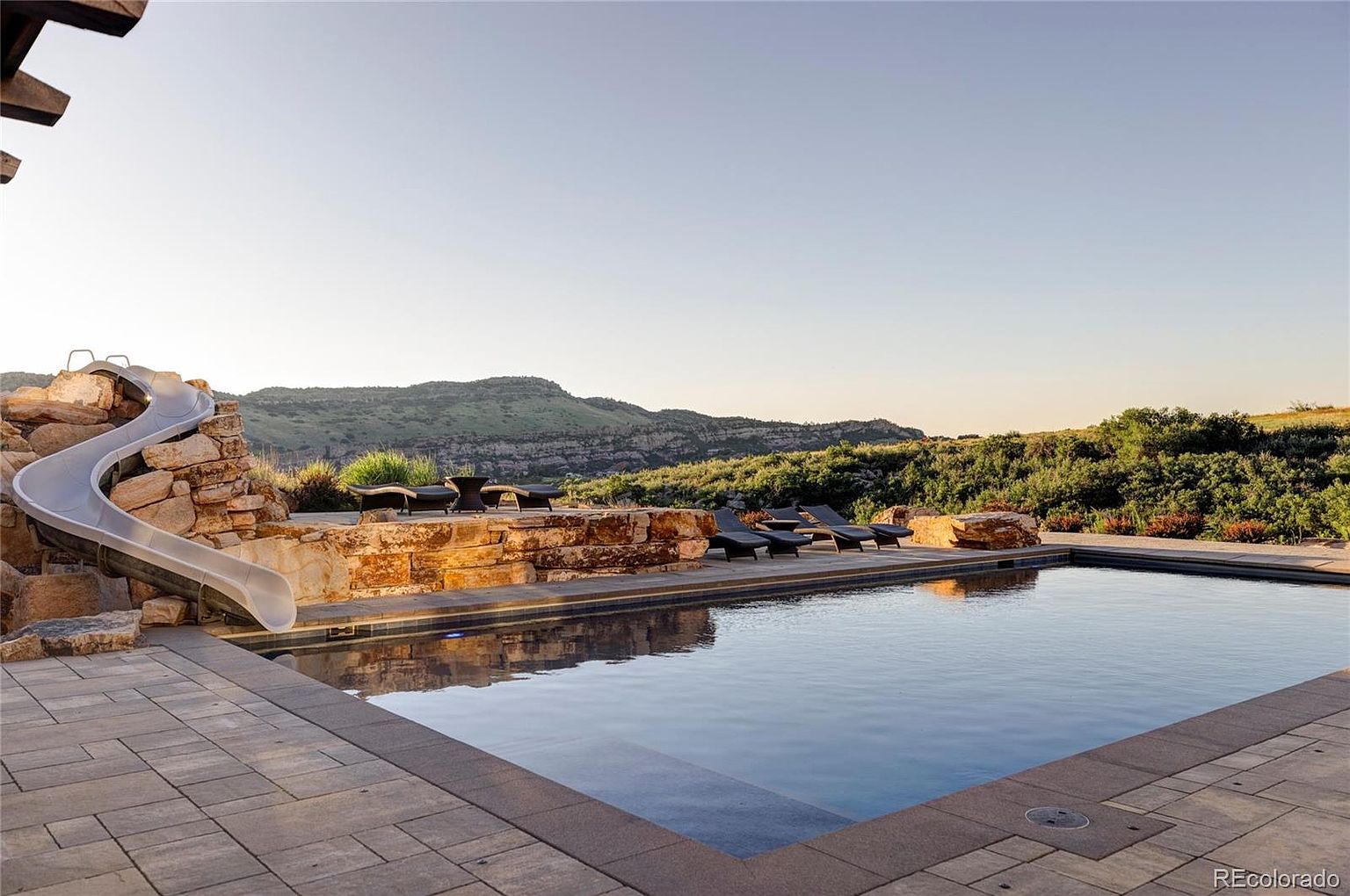 This image showcases a luxurious outdoor pool area with a slide built into a rock formation. The pool is surrounded by a stone patio and lounge chairs, with a scenic mountain view in the background. The setting evokes a sense of relaxation and high-end outdoor living.